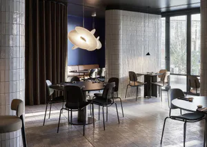 Modern restaurant interior with dark round tables, upholstered chairs, large sculptural white pendant light, blue accent wall, white tiled columns, and patterned terrazzo floor.