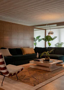Modern living room interior with dark green velvet sectional sofa, striped lounge chair, light wood coffee table, patterned area rug, wood-paneled walls and ceiling, and large potted plants.