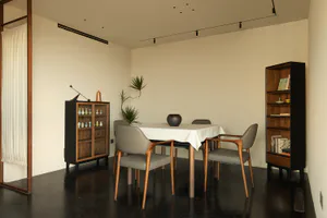 Modern minimalist dining room with a rectangular table covered in white linen, four grey upholstered wooden dining chairs, a tall dark wood display cabinet filled with ceramics, a matching tall bookshelf, potted plants, and a dark glossy floor under track lighting.