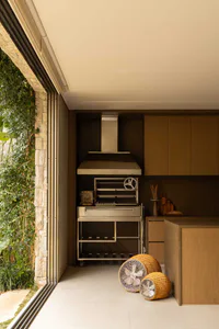 Modern outdoor kitchen with a stainless steel pizza oven, range hood, wooden cabinets, and two woven floor fans placed on light-colored tile flooring next to a stone wall and large glass doors.