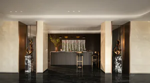 Modern luxury kitchen bar area with dark marble floor, beige marble walls, bronze sculptures on marble pedestals, and a central bar with gold bar stools under pendant lights.