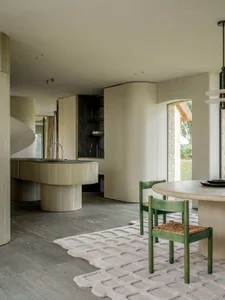 Modern minimalist dining area with curved oak kitchen island, moss green wooden chairs with woven seats, and a sculptural white rug on grey tiled floor.