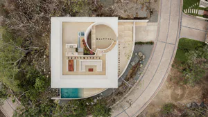 Aerial photograph of a modern minimalist villa with white concrete walls, a large circular open-air courtyard with stone steps, a rectangular swimming pool, and surrounding tropical vegetation.