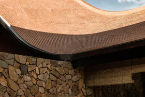 Close-up view of a curved terracotta tile roof with deep shadows, meeting a rustic fieldstone wall and bamboo blind below.