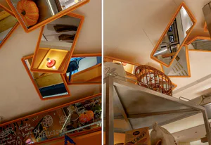 Low-angle view of irregularly shaped orange-framed mirrors mounted on a cafe ceiling, reflecting baked goods, pumpkins, and warm lighting.