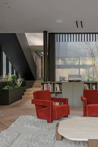 Modern living room featuring two red velvet armchairs on a white shaggy rug, a marble coffee table, built-in planter with cacti, wooden staircase, and large glass windows overlooking trees.