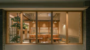 Exterior night view through large wooden-framed glass windows into a warmly lit Japanese restaurant featuring oak furniture, wooden counters, pendant lights, and indoor plants.