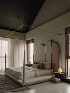 Modern minimalist bedroom featuring a tall brass four-poster bed with textured fabric canopy, layered neutral bedding, arched woven room divider, and dark ceiling with dramatic lighting.