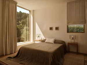 Minimalist bedroom with brown linen bedding, wooden nightstands, large windows with beige curtains, and woven jute rug, bathed in natural morning light.