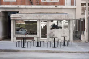 Modern minimalist concrete cafe with large glass windows, black round high tables and stools arranged on a light gray tiled pavement, with a blurred person walking past.