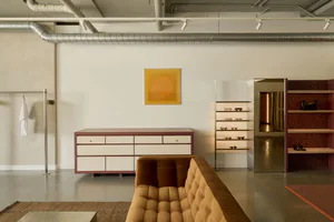 Modern boutique interior with a mustard tufted sofa in the foreground, a pink-trimmed white credenza, illuminated eyewear display shelves, and a large ochre circular artwork on a white wall with exposed silver ductwork.