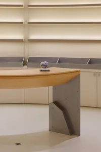 Modern curved reception desk made of light oak wood with a thick angular steel base, displaying a purple amethyst mineral specimen on top, set against minimalist beige walls with floating shelves and soft backlighting.