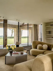 Modern organic living room featuring a curved mustard-yellow bouclé sofa, metallic coffee table, large limestone walls, and expansive glass doors opening to a green landscape.