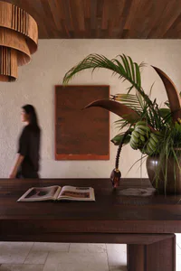 A blurred woman walks past a dark wooden console table featuring an open book, a large potted banana plant with green fruit, and a hanging wooden pendant lamp against a textured wall with a rust-colored artwork.