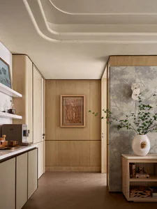 Modern minimalist entryway with light oak built-in cabinetry, marble countertops, sculptural wavy ceiling, framed forest artwork, and a console table with botanical wallpaper and a large white vase with green branches.