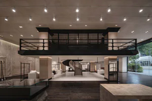 Interior view of a luxury fashion boutique with a dramatic double-height ceiling, black metal railing on the mezzanine, a sculptural spiral staircase in the center, travertine stone walls and floors, and minimalist white seating areas.
