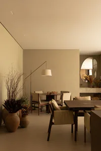 Modern minimalist dining room featuring a round walnut chess table with upholstered chairs, a dramatic floor lamp, large terracotta vases with dried branches, and an oval mirror reflecting another part of the home.