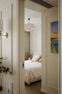 View through a doorway into a serene modern classic bedroom with neutral bedding, layered pillows, pendant light, and a colorful road landscape painting on the wall.