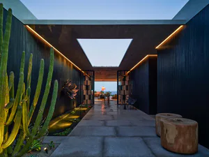 Modern outdoor corridor with charred wood walls, linear LED lighting, tall cactus garden, water channel, and open sky light overlooking the sea at dusk.