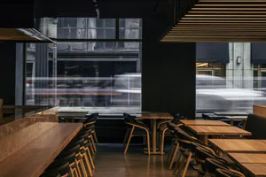 Modern restaurant interior featuring dark oak tables and chairs, black upholstery, dramatic linear ceiling lights, and large windows showing motion-blurred traffic outside.