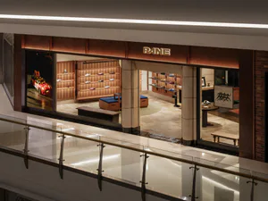 High-angle view of a luxury shoe store named PANE with warm wooden shelving, blue ottomans, stone flooring, and a large red race car photograph on the left wall.