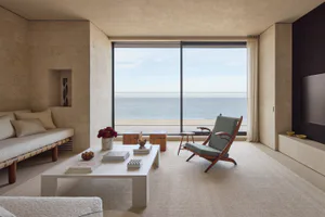 Modern minimalist living room with travertine walls, a built-in daybed, white coffee table, and a wooden lounge chair facing a large glass window overlooking the ocean and beach.