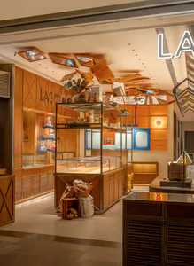 Modern bakery interior showcasing warm wooden cabinetry, glass display cases, and a creative ceiling installation of suspended orange-tinted mirrors reflecting warm lighting.