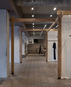Long perspective view of a minimalist boutique interior featuring exposed concrete walls, large wooden structural beams, tiled floor, and clothing displays under track lighting.
