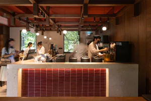 Modern cafe interior featuring a terrazzo counter with red tiled front, warm wood elements, and staff preparing drinks.