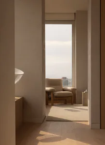 View from a doorway into a minimalist room featuring a beige upholstered oak armchair, a matching ottoman, and a large window overlooking a hazy city skyline with dramatic morning light casting shadows on light wood floors.