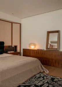 Modern minimalist bedroom interior with a walnut dresser, large framed mirror, glowing Murano-style glass lamp, neutral linen bed, and black and white patterned rug.
