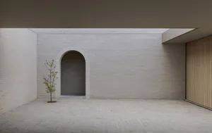 Minimalist interior courtyard with white brick walls, a central arched doorway, a small leafy tree, and a wooden slat wall on the right under an overcast sky.