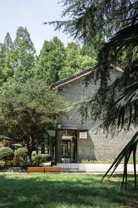 傳統灰磚古蹟建築外觀，庭院入口被茂密綠樹環繞，前景有木製長椅。