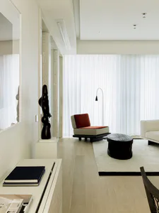 Modern minimalist living room featuring a tall abstract dark wood sculpture on a white pedestal, a red and beige lounge chair, a dark wood coffee table on a light rug, sheer white curtains, and light oak flooring.