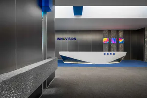 Modern corporate reception area featuring a white geometric reception desk with blue metallic base, stainless steel walls, blue carpet, and colorful digital art displays on the back wall with the company name INNOVISION.