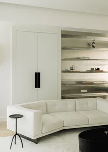 Modern minimalist living room with a large white curved sectional sofa, black round coffee table, black tripod side table, and built-in white oak cabinetry with floating stainless steel shelves displaying books and decorative objects.