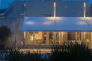 現代混凝土建築，大型玻璃門通往設有白色桌椅的戶外用餐露台，由長型白色遮陽棚與垂直燈具在黃昏時分照亮，前景有高聳的草叢。