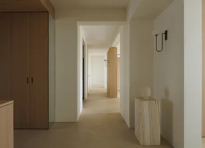 Long minimalist hallway featuring a travertine stone pedestal with a white ceramic vase, warm wood cabinetry, black wall sconce, and light oak flooring.