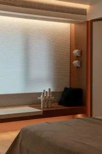 Modern minimalist bedroom interior with a wooden window bench, white abstract sculpture, black cushion, pleated blinds, and warm orange accent wall.
