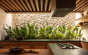 Modern kitchen interior with a large natural stone wall covered in lush tropical plants, wooden ceiling slats, dark stone countertops, and a gas cooktop on a kitchen island.