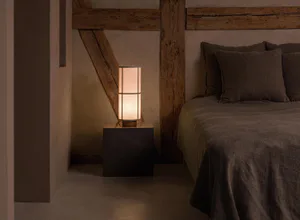 Minimalist bedroom corner with a dark marble cube bedside table holding a lit rectangular linen table lamp, set against a textured plaster wall with exposed reclaimed wooden beams and a bed dressed in earthy brown linen bedding.