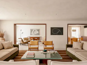 Modern living room with two green velvet sofas facing each other, two mustard leather teak armchairs, a glass coffee table with books, and views into a dining area and secondary seating space.