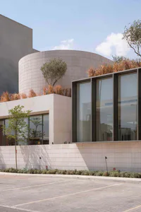 Modern curved building with travertine stone cladding, large glass windows, and a rooftop garden featuring olive trees and ornamental grasses, set against a clear sky with a paved foreground.