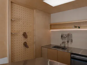 Modern minimalist kitchen interior with light birch plywood pegboard wall holding two brown aprons, stainless steel double sink, wooden upper shelf with integrated lighting, and wooden cabinetry.