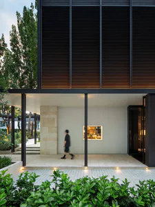 Modern minimalist courtyard of a contemporary house featuring a large travertine stone wall, dark horizontal wooden louvers with warm backlighting, a person walking across the paved area, and lush green foreground planting.