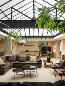 Modern industrial-style lounge area featuring a large brown leather sectional sofa, round glass coffee tables, wooden armchairs, abundant indoor plants, and a striking black-framed glass skylight ceiling with a chrome pendant lamp.