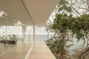 Modern minimalist house interior with floor-to-ceiling glass walls overlooking the ocean, featuring a travertine stone terrace and lush tropical trees on a cliffside location.