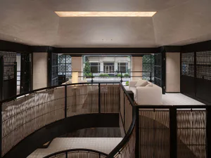 Modern mezzanine living area featuring curved dark wood railing with vertical wood screens, white L-shaped sofa, textured beige walls, and a skylight above, overlooking a courtyard garden.