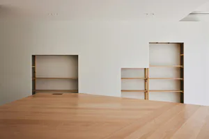 Modern minimalist interior featuring light oak built-in shelving units recessed into white walls, with a large light oak wooden table in the foreground.