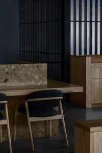 Modern dining area featuring a thick terrazzo-topped table with solid oak legs, paired with oak-framed dining chairs upholstered in dark navy fabric, set against a dramatic black slatted wall.